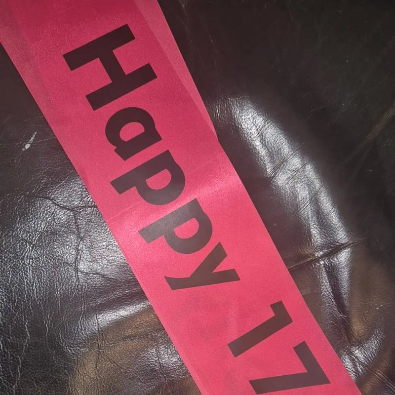 A pink sash with the words 'Happy 17' printed in black text, laid diagonally across a dark brown leather surface.