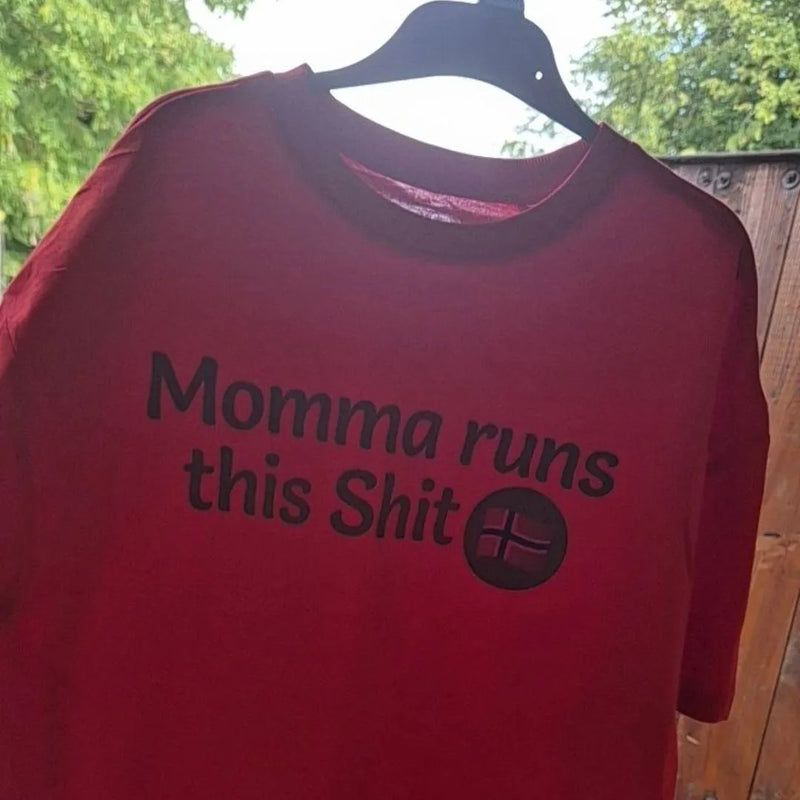 A red short-sleeve t-shirt hanging on a black hanger outdoors, featuring the black printed text 'Momma runs this' alongside a small black flag graphic.