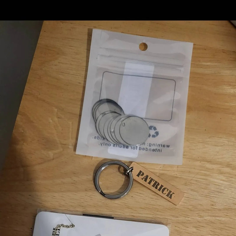 A small metal keychain with a rectangular wooden tag engraved with the name 'PATRICK' lies on a light wood tabletop next to a clear plastic bag containing several round silver keychain discs.