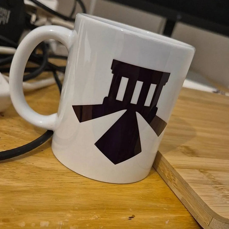 A white ceramic coffee mug with a black geometric graphic of a classical building, resting on a wooden cutting board with a blurred desk background.