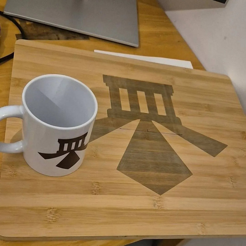A white ceramic mug with a black geometric logo sits on a light wood tray or placemat featuring a dark printed graphic of a classical building and geometric shapes, placed on a wooden desk next to a laptop.