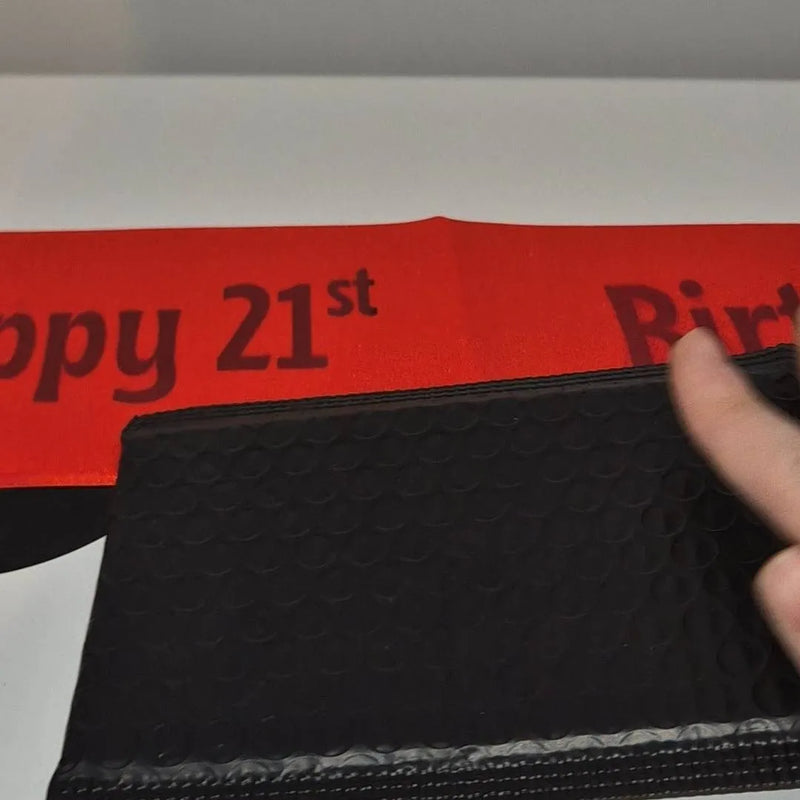 A close-up of a black textured pouch or sleeve with a finger touching it, placed in front of a red banner printed with 'Happy 21st Birthday' in dark lettering.