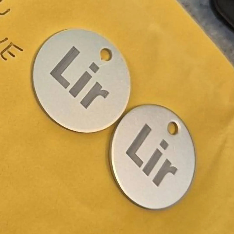 Two round silver metal tags with a small hole at the top and the engraved text 'Lir', placed on a yellow surface.