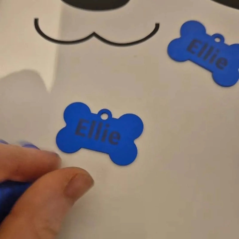 A close-up of a hand placing a blue bone-shaped pet name tag labeled 'Ellie' onto a white surface, with two matching tags visible.